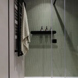 Nowoczesna łazienka z kabiną prysznicową ze szklanymi drzwiami, zielonymi, pionowo żłobionymi płytkami, czarnym grzejnikiem drabinkowym z zawieszonym ręcznikiem w paski oraz półką z trzema...