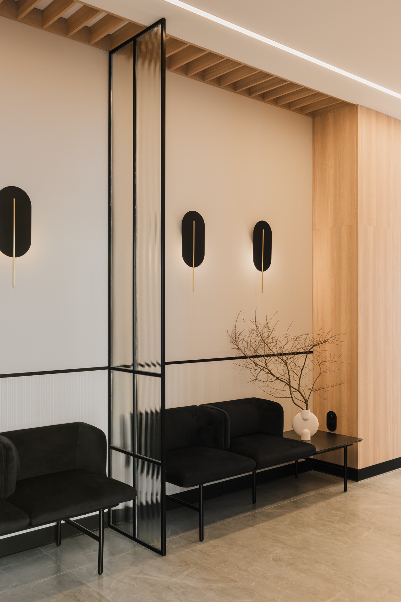 Wnętrze poczekalni z czarnymi sofami, szklaną ścianką działową z czarną ramą, minimalistycznymi lampami ściennymi i dekoracyjną gałązką w białym wazonie.