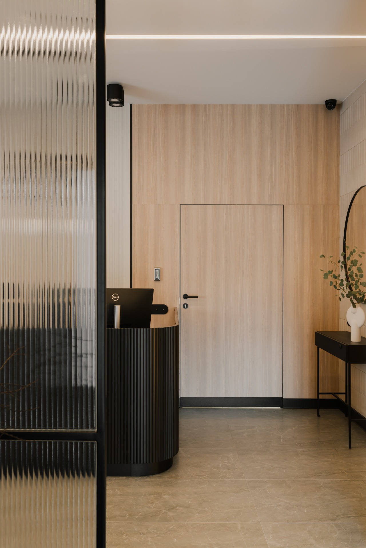 Nowoczesne biuro: recepcja z czarnym biurkiem recepcyjnym, laptopem Dell, ścianą wyłożoną drewnem i ukrytymi drzwiami, oświetleniem LED oraz dekoracyjnym lustrem z roślinami.