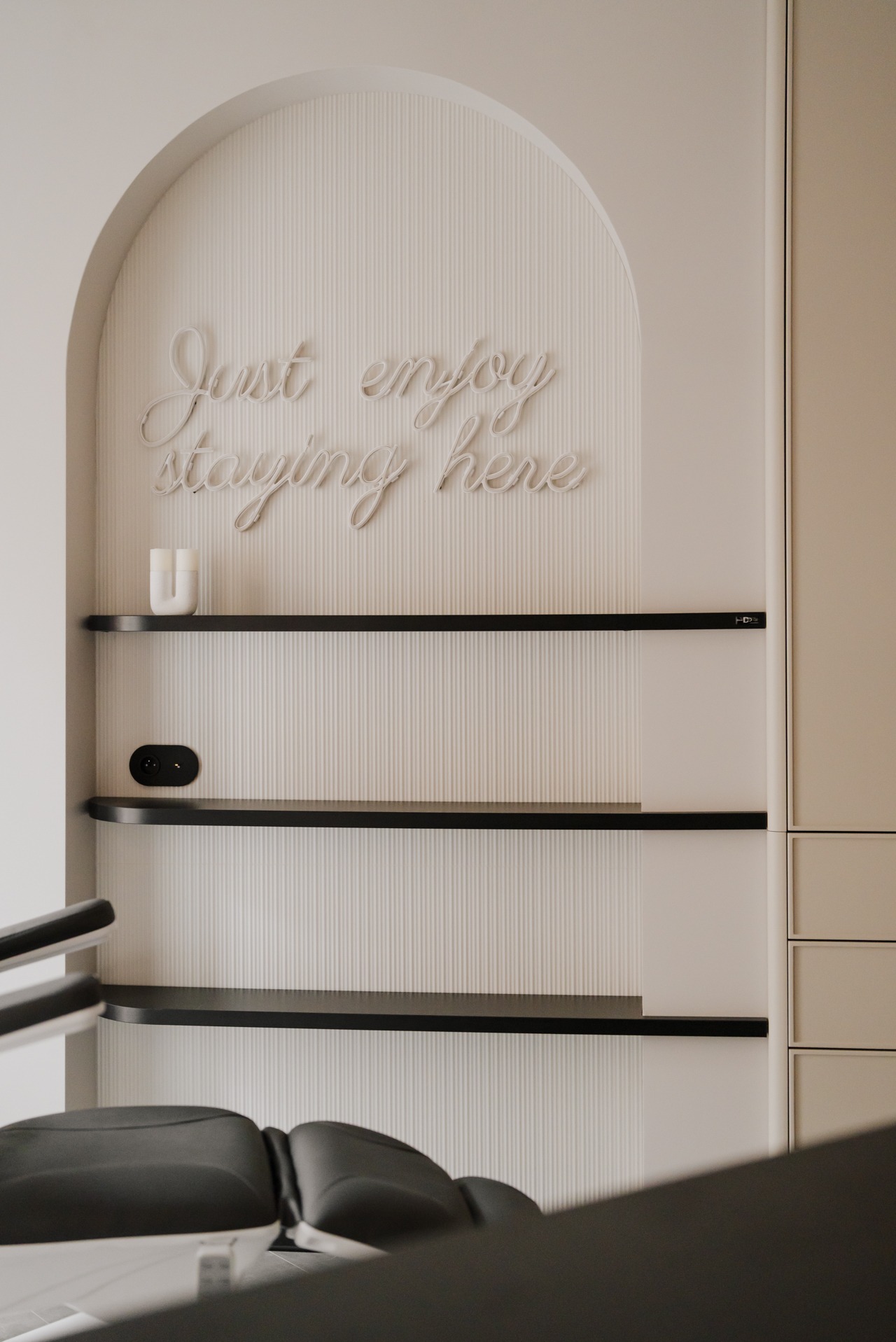 Kremowe wnętrze gabinetu z półkami i neonowym napisem 'Just enjoy staying here' nad fotelem zabiegowym.
