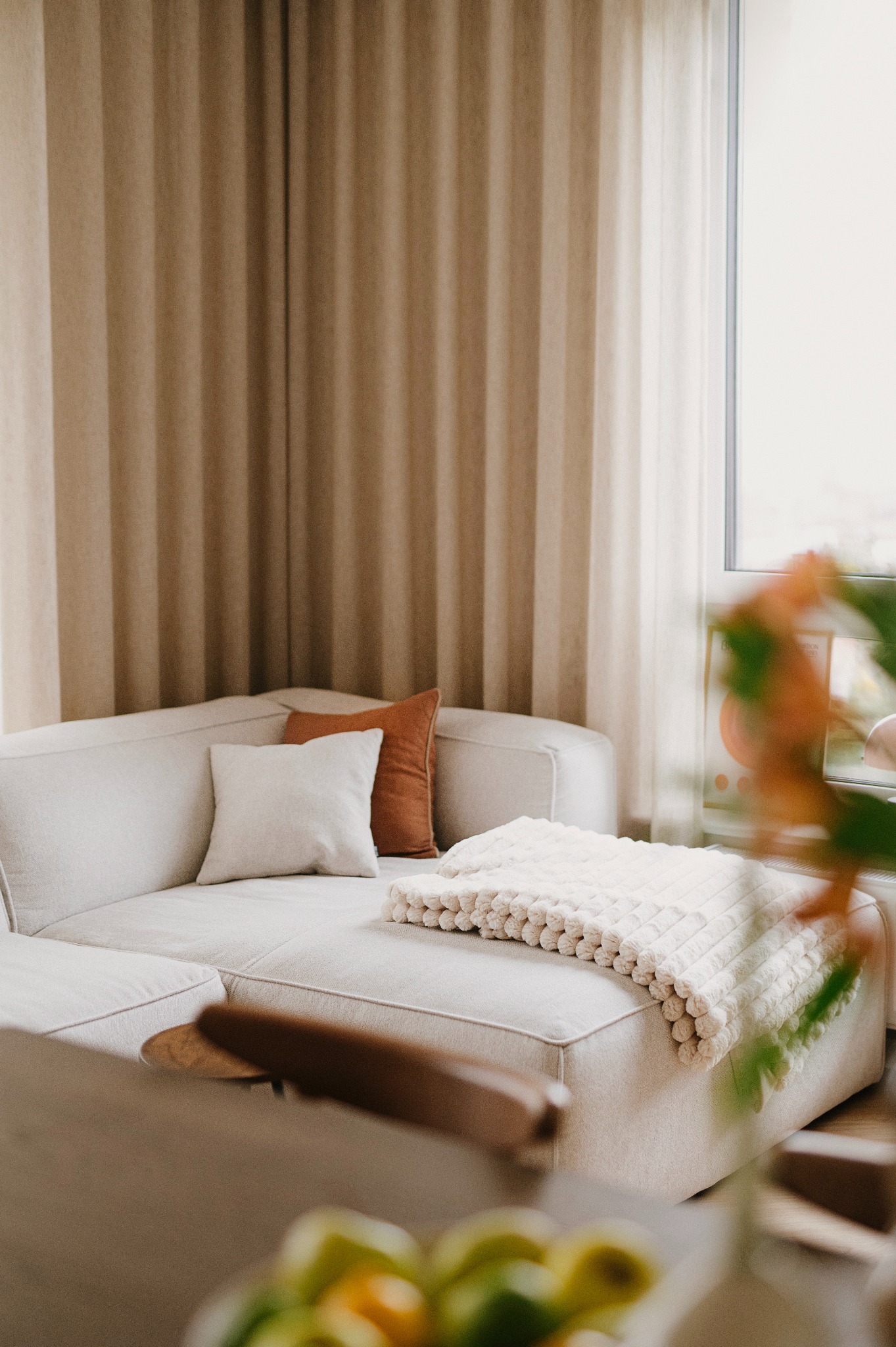 Jasny narożnik z poduszkami w odcieniach beżu i brązu, przykryty kremowym pledem o wyraźnej fakturze, na tle okna z beżowymi zasłonami.