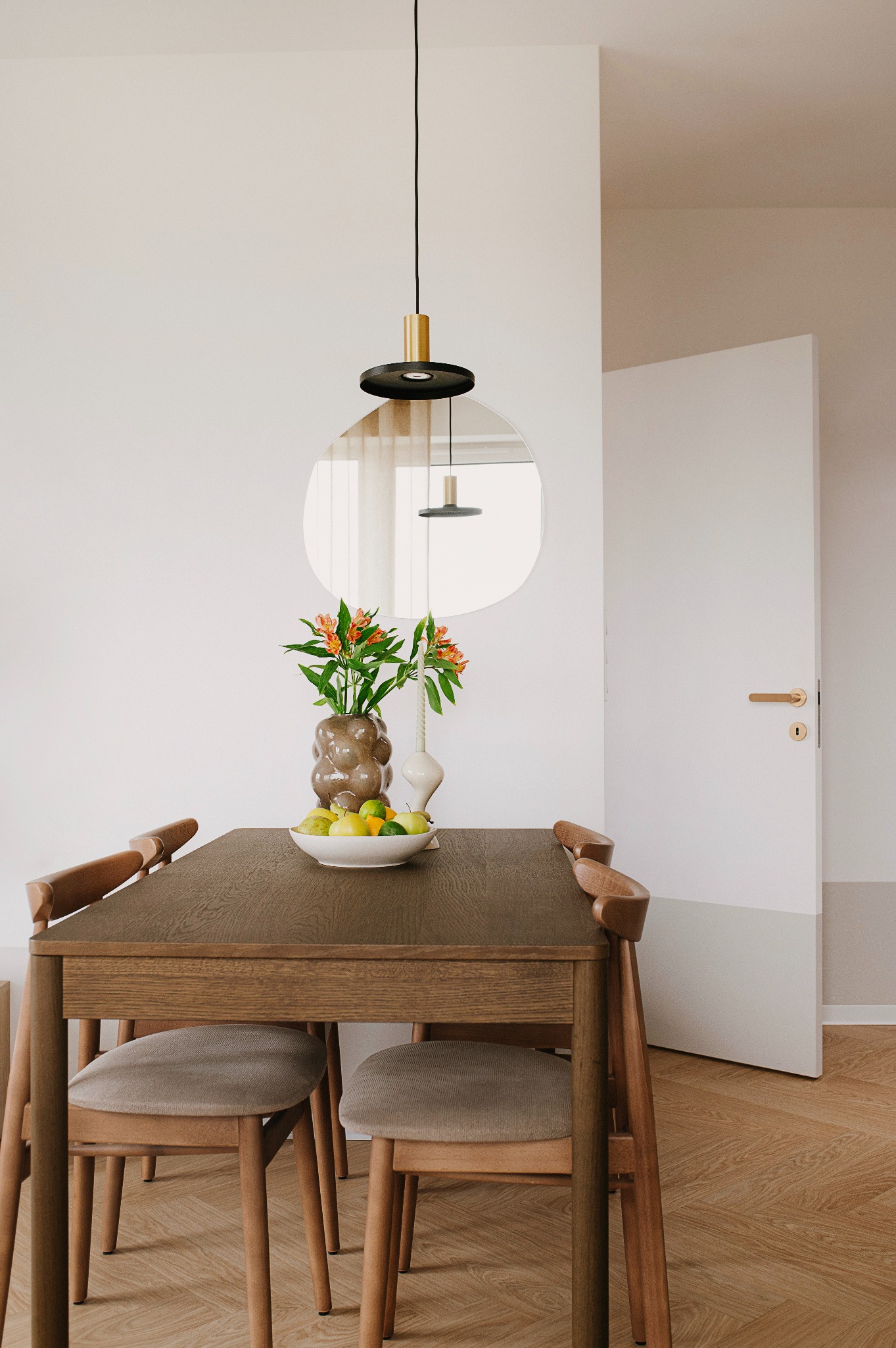 Nowoczesna jadalnia z drewnianym stołem, krzesłami z szarą tapicerką, wazonem z pomarańczowymi kwiatami i owocami, oświetlona designerską lampą wiszącą i odbijająca się w okrągłym lustrze na ścianie.