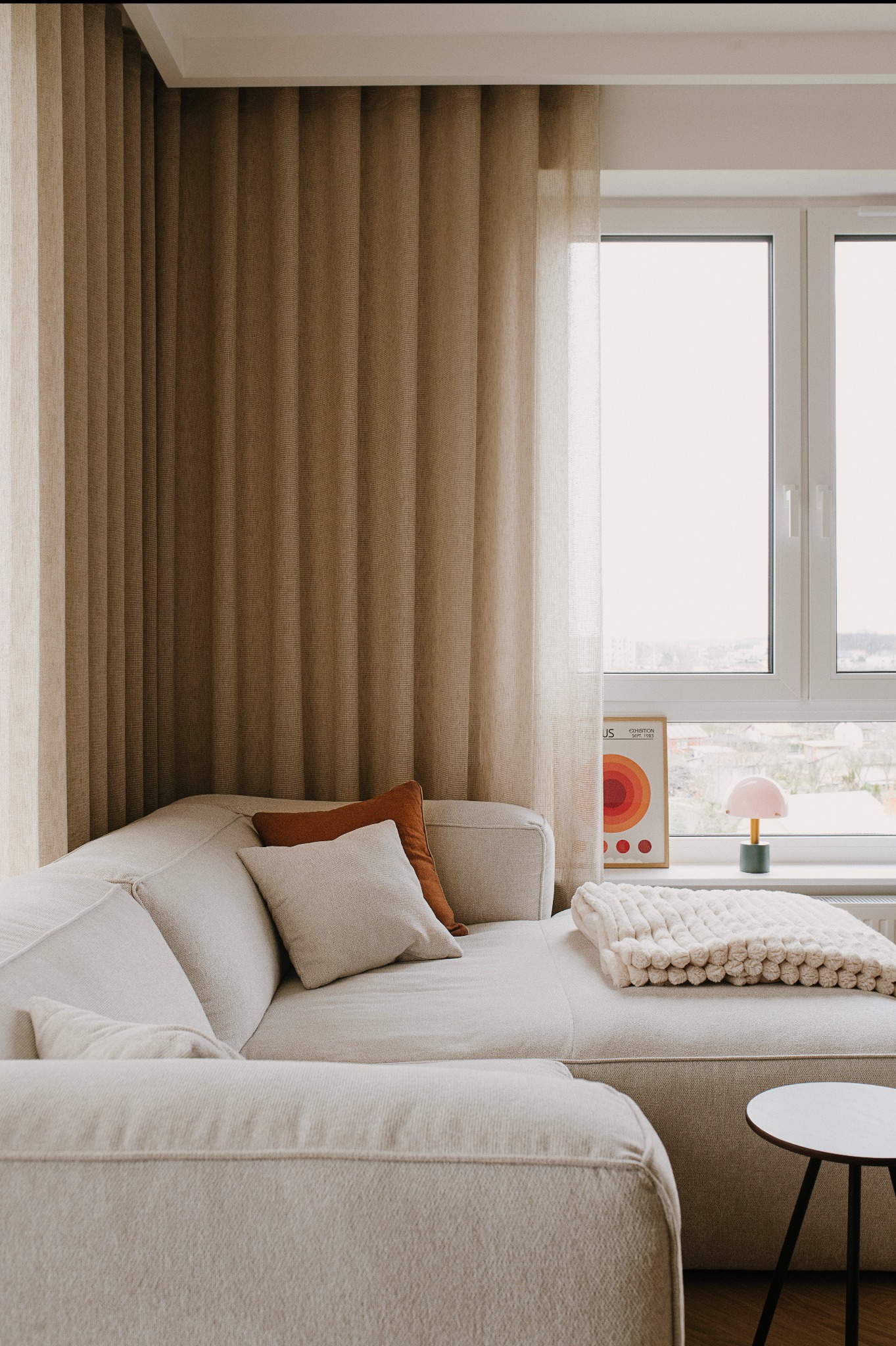 Jasna, narożna sofa z poduszkami i kocem w neutralnych odcieniach, ustawiona przy oknie z plakatem i lampką, obok mały stolik kawowy.