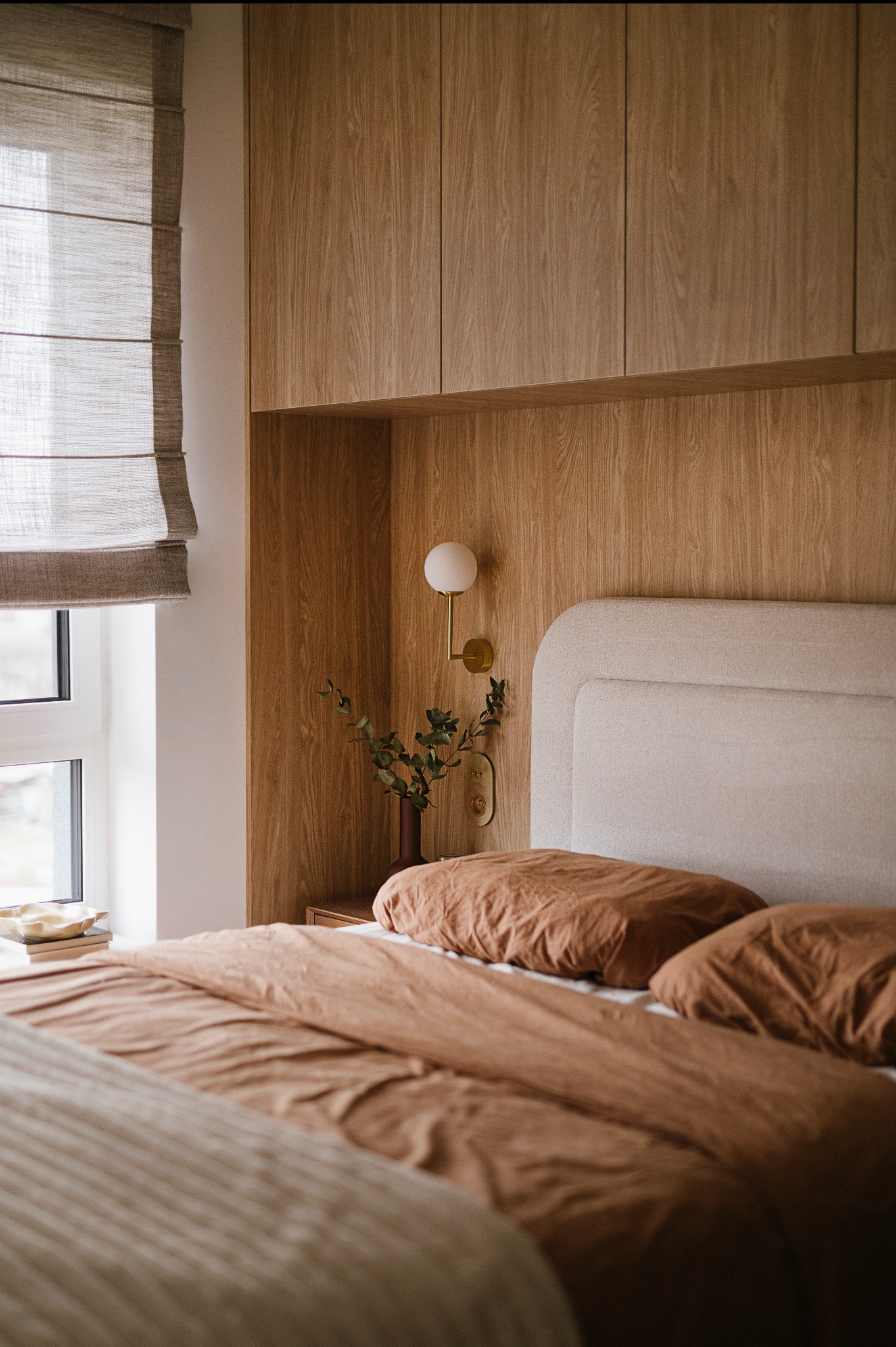 Sypialnia w ciepłych beżach i brązach: drewniana zabudowa nad łóżkiem, minimalistyczna lampka ścienna, lniana pościel w odcieniu terakoty, roleta rzymska przy oknie.