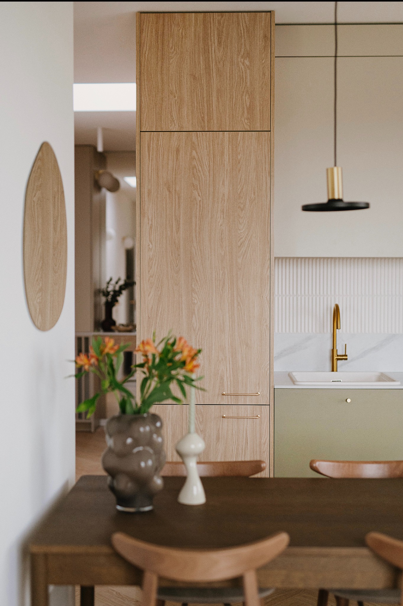 Nowoczesna kuchnia z drewnianymi frontami szafek, złotą armaturą i minimalistyczną lampą wiszącą, stół z wazonem pomarańczowych kwiatów na pierwszym planie.
