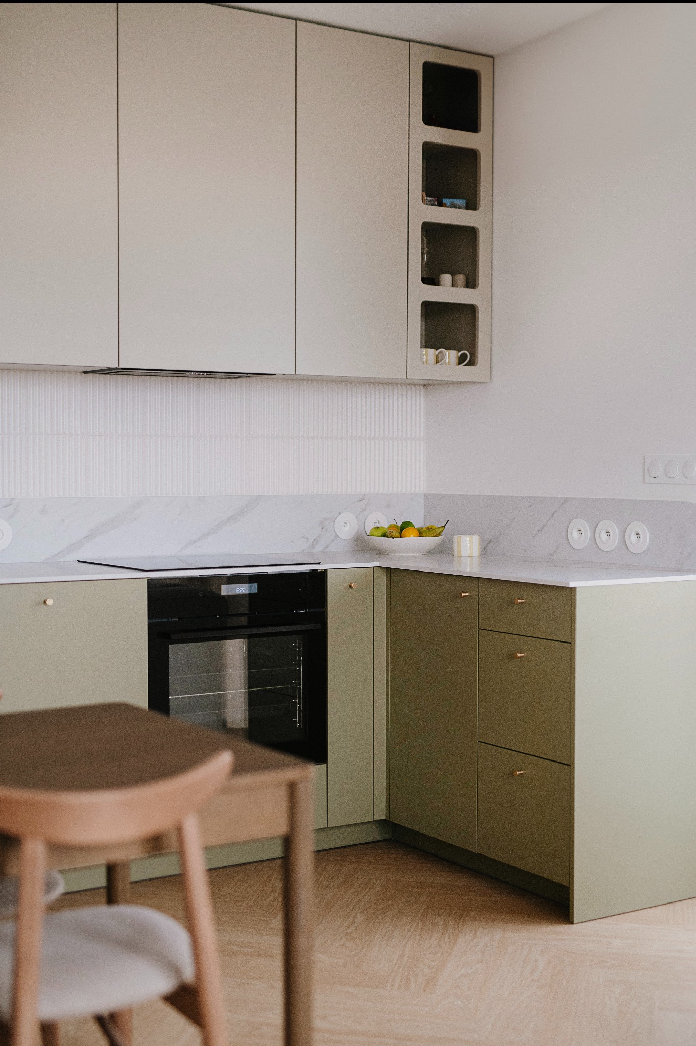 Nowoczesna kuchnia z oliwkowymi szafkami, marmurowym blatem i drewnianą podłogą; na blacie misa z owocami i ceramiczne gniazdka elektryczne.