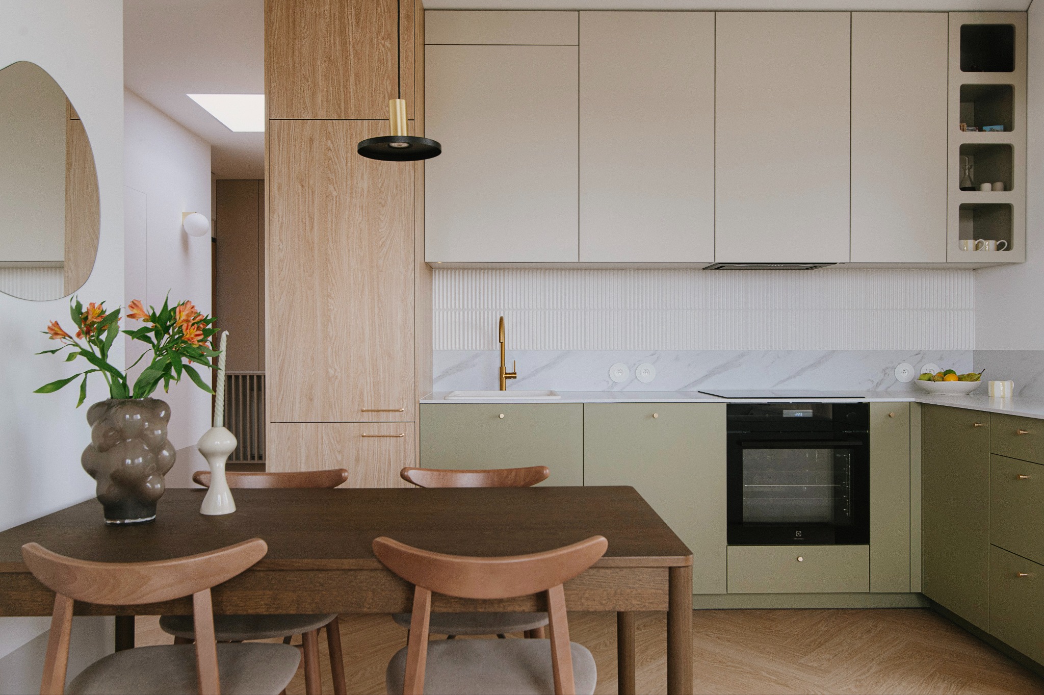 Nowoczesna kuchnia z drewnianym stołem i krzesłami, kwiaty w ozdobnym wazonie na blacie, minimalistyczne szafki w odcieniach beżu i zieleni, złota armatura i lampa wisząca.