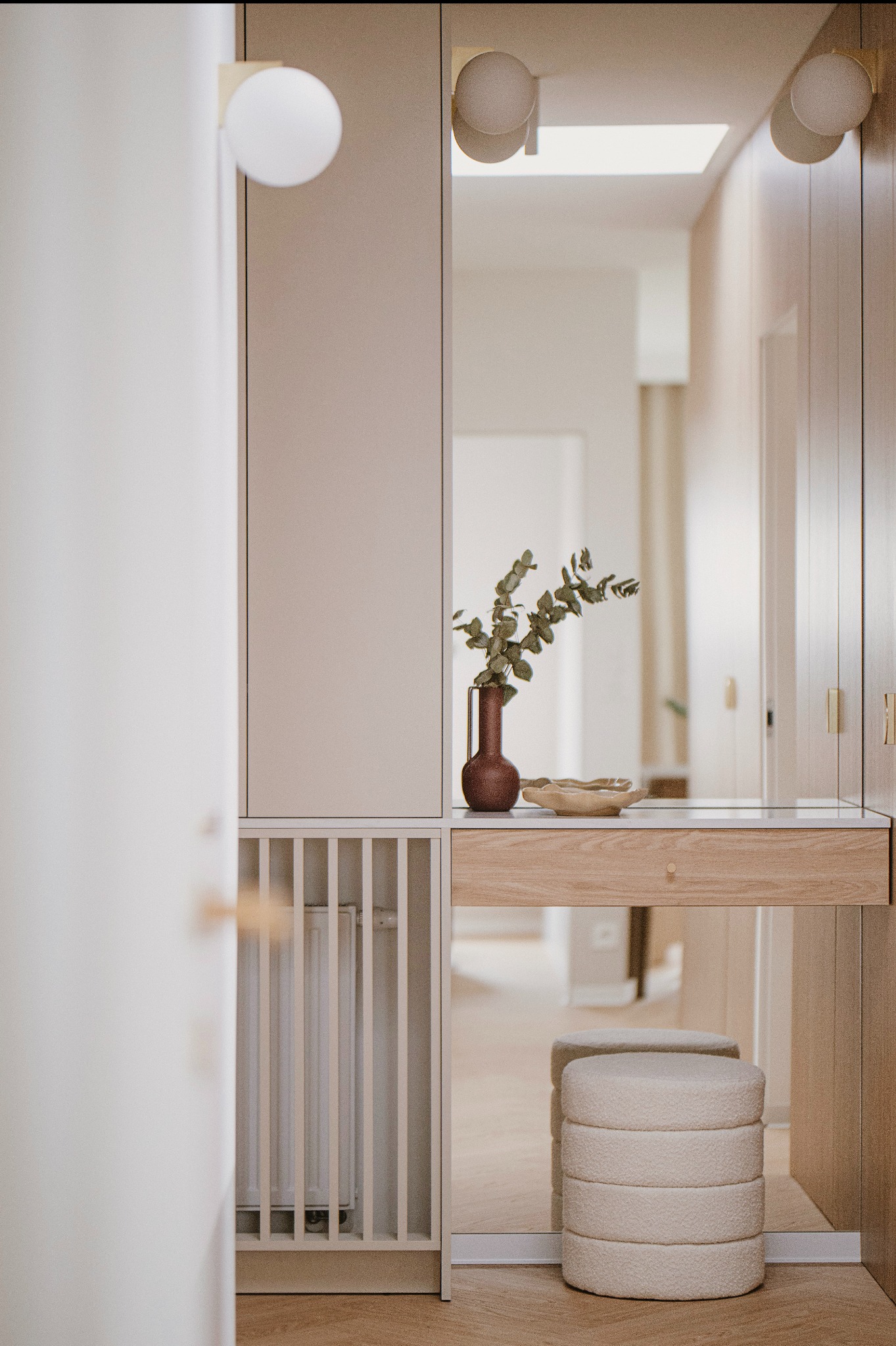 Jasne wnętrze przedpokoju z toaletką, lustrem, pufą i dekoracyjnym wazonem z eukaliptusem, oświetlone lampami kulami