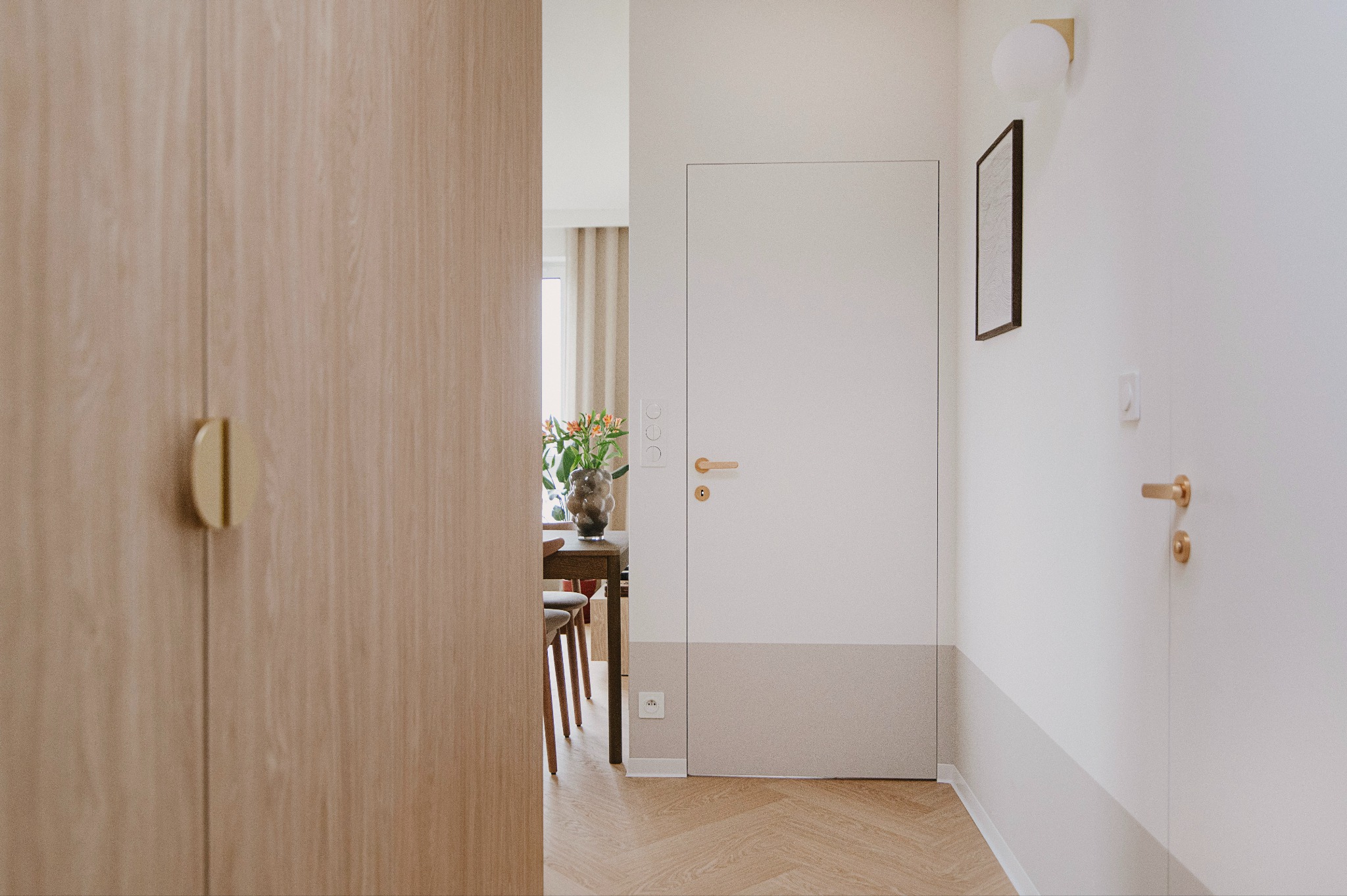 Minimalistyczny przedpokój z widokiem na jadalnię, parkiet w jodełkę, białe ściany z szarym pasem, zamknięte drzwi z złotą klamką, szafa z drewnianymi frontami i złotymi uchwytami.