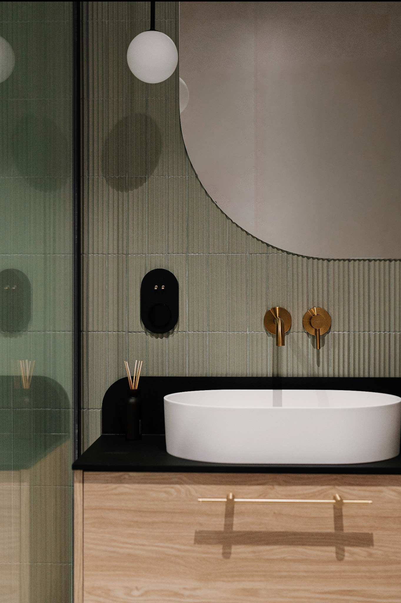 Nowoczesna łazienka z owalną umywalką nablatową, złotą armaturą, zielonymi płytkami ściennymi z subtelnym wzorem i okrągłym lustrem bez ramy.