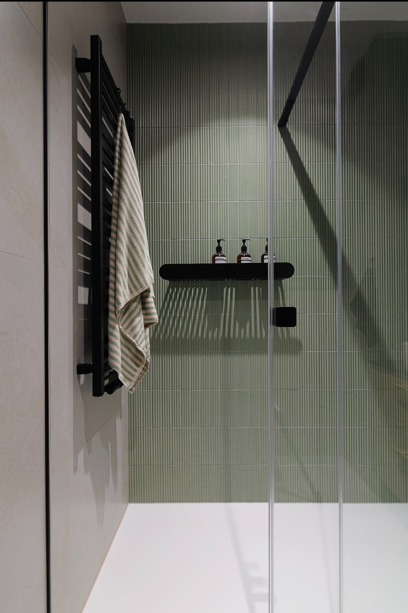 Nowoczesna łazienka z kabiną prysznicową ze szklanymi drzwiami, zielonymi, pionowo żłobionymi płytkami, czarnym grzejnikiem drabinkowym z zawieszonym ręcznikiem w paski oraz półką z trzema...