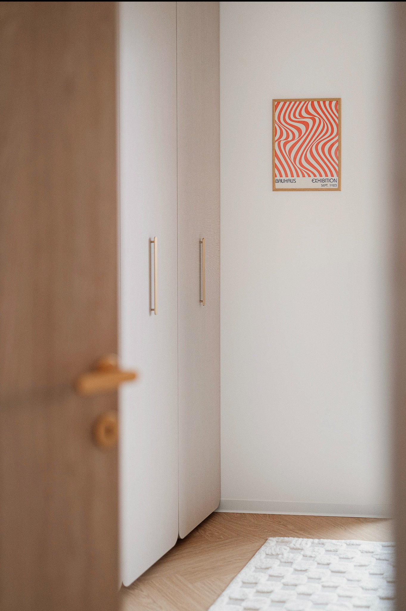Minimalistyczna szafa wnękowa z długimi, złotymi uchwytami, uzupełniona plakatem Bauhaus na białej ścianie i wzorzystym dywanem na podłodze z parkietu ułożonego w jodełkę.