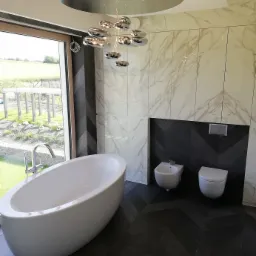 Nowoczesna łazienka z wolnostojącą białą wanną, bidetem i toaletą, marmurowe płytki na ścianach i podłoga ułożona w jodełkę, designerska lampa sufitowa.