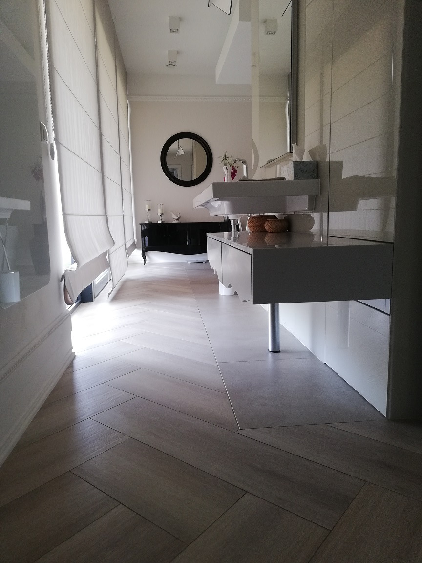 Nowoczesna łazienka z podłogą ułożoną w jodełkę, białą szafką pod umywalką na metalowej nodze, dużym lustrem i roletami rzymskimi w oknach.