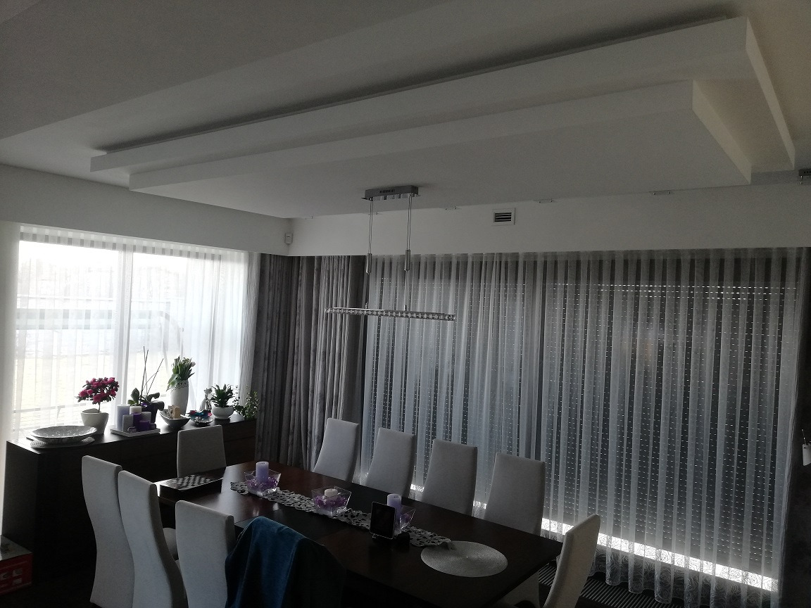 Nowoczesna jadalnia z dużym oknem, zasłoniętym przezroczystymi firanami i szarymi zasłonami, elegancki stół z dekoracjami, białe krzesła i podwieszany sufit z oświetleniem.