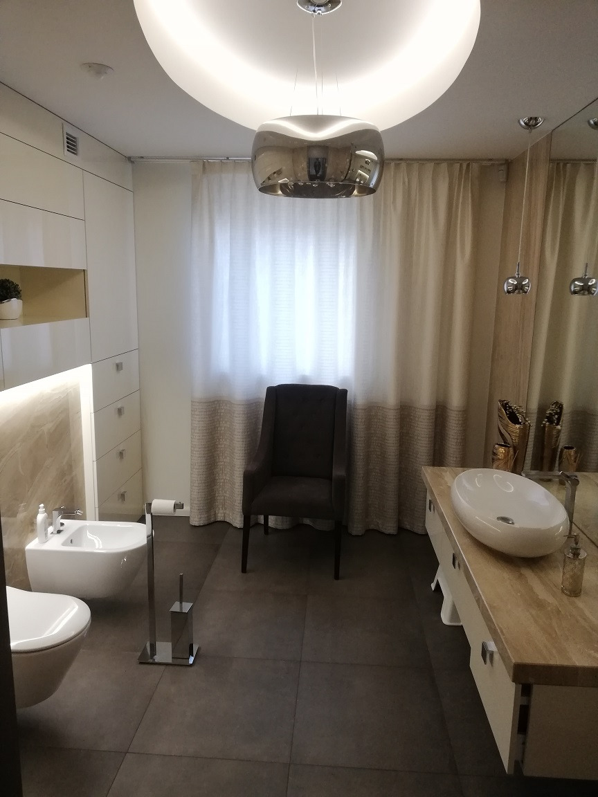 Nowoczesna łazienka z ciemnymi płytkami podłogowymi, białymi sanitariatami, kremowymi zasłonami i designerską lampą wiszącą.