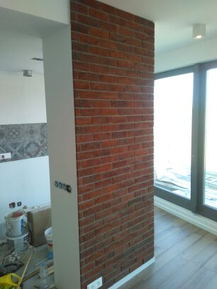 Ściana wykończona czerwoną cegłą dekoracyjną, połączona z białą kolumną w nowoczesnym wnętrzu z widocznymi puszkami elektrycznymi i elementami wykończeniowymi na podłodze.