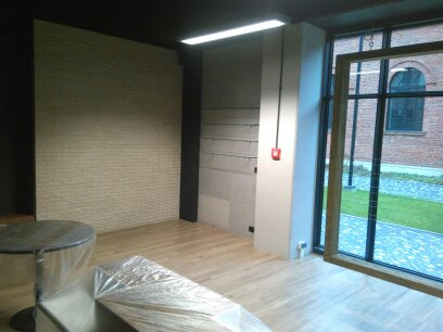 Wnętrze pomieszczenia z wykończoną ścianą z cegieł, drewnianą podłogą i dużym oknem z widokiem na zewnątrz, meble zabezpieczone folią ochronną.