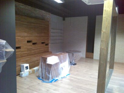 Wnętrze w trakcie remontu: drewniana okładzina ściany z dekoracyjnymi wnękami, podłoga z jasnego drewna, meble zabezpieczone folią, ściana z białej cegły w tle.