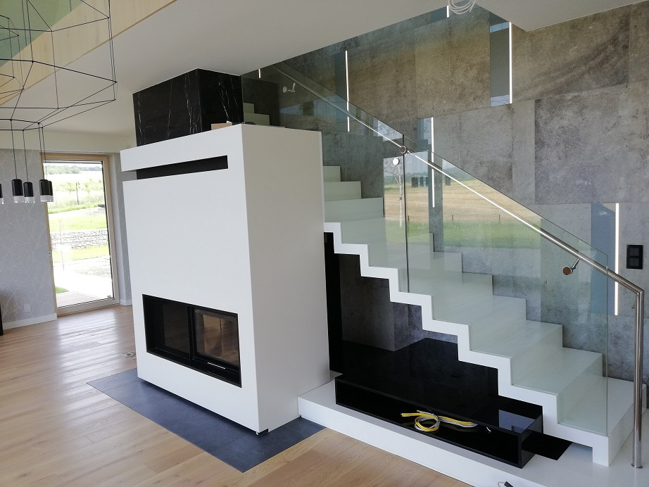 Nowoczesny salon z białymi schodami z szklaną balustradą, minimalistycznym kominkiem obudowanym białą płytą i podłogą z jasnego drewna.