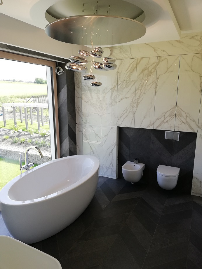Nowoczesna łazienka z wolnostojącą białą wanną, bidetem i toaletą, marmurowe płytki na ścianach i podłoga ułożona w jodełkę, designerska lampa sufitowa.
