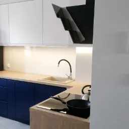 Nowoczesna kuchnia z granatowymi szafkami dolnymi, jasnym drewnianym blatem i białymi szafkami górnymi, minimalistyczny okap, płyta indukcyjna z patelnią i czajnikiem.