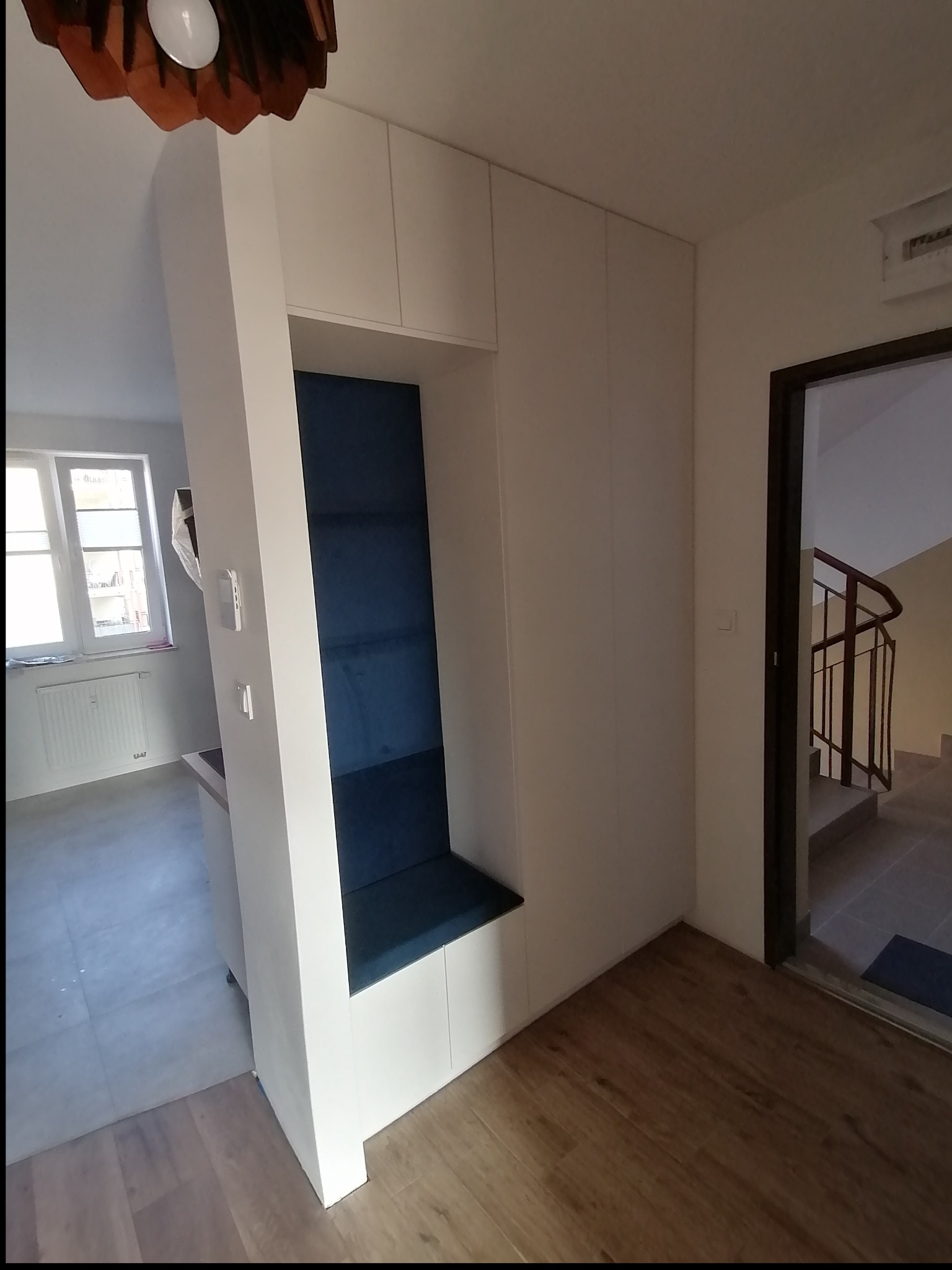 Biała zabudowa wnęki z siedziskiem tapicerowanym na granatowo, wkomponowana w przedpokój z drewnianą podłogą i widokiem na klatkę schodową.