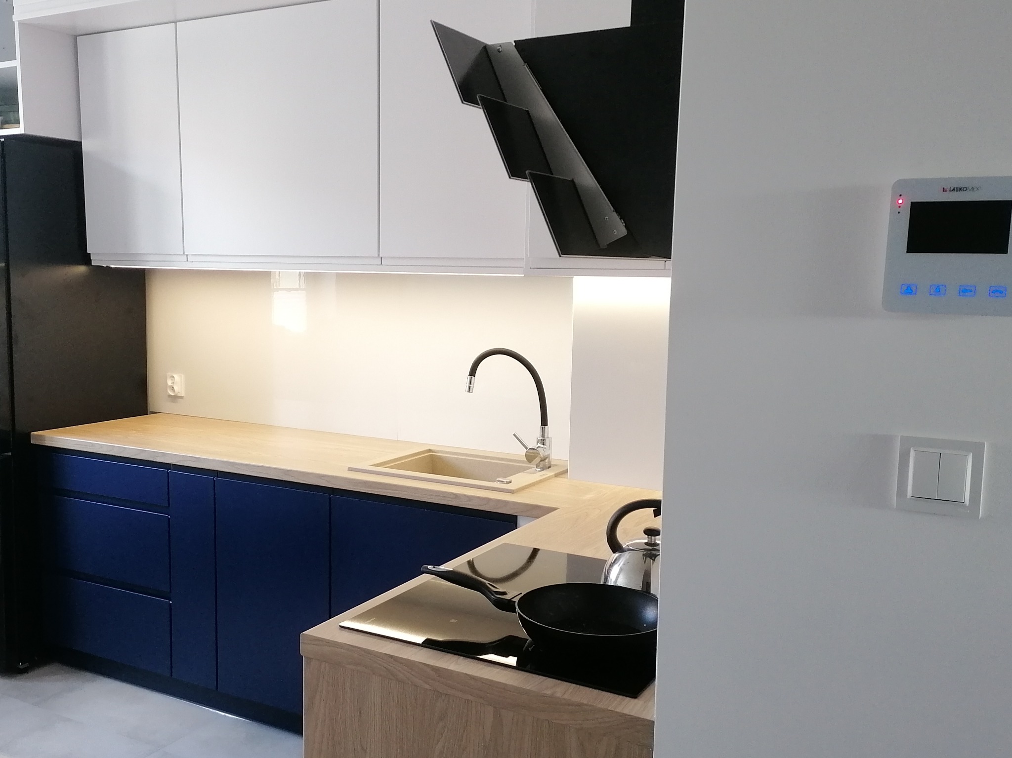 Nowoczesna kuchnia z granatowymi szafkami dolnymi, jasnym drewnianym blatem i białymi szafkami górnymi, minimalistyczny okap, płyta indukcyjna z patelnią i czajnikiem.