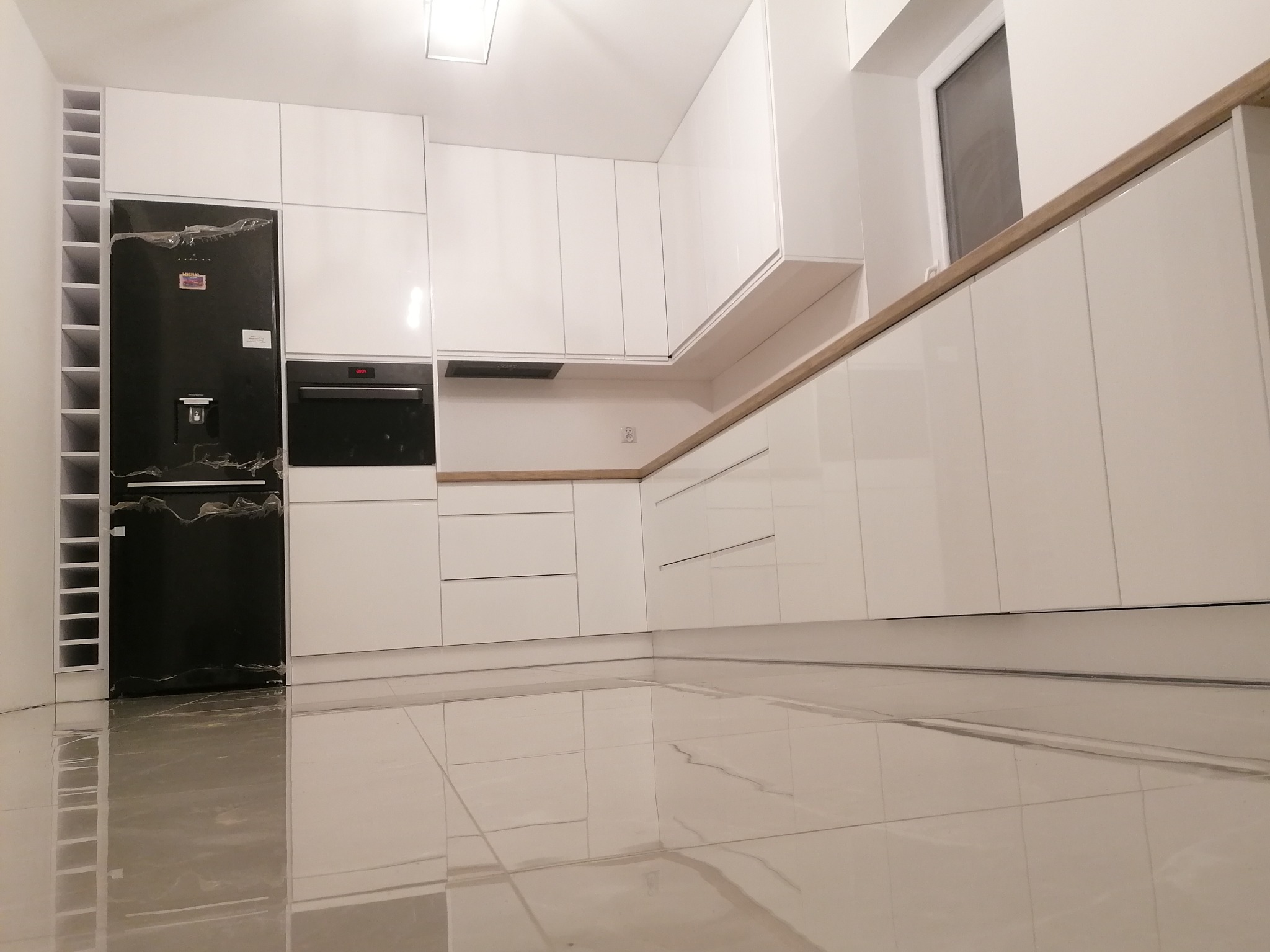 Nowoczesna, minimalistyczna kuchnia z białymi, lakierowanymi frontami, drewnianym blatem i czarną lodówką z kostkarką, widok z niskiego kąta ukazujący połyskliwe płytki podłogowe.