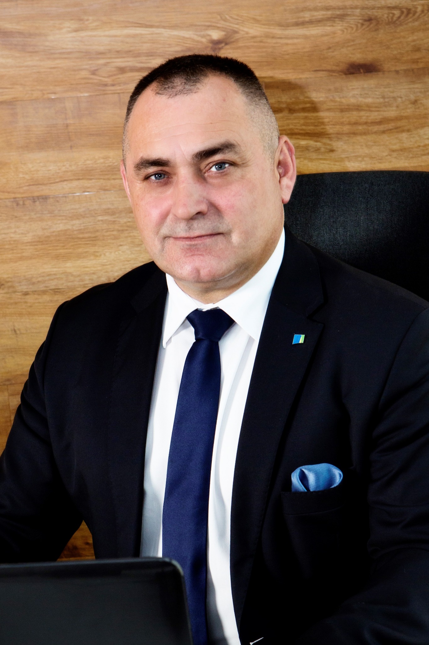 Portret mężczyzny w garniturze z niebieskim krawatem i poszetką, siedzącego przy biurku z drewnianym tłem, z przypinką w barwach flagi Ukrainy na klapie.