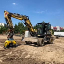 Żółta koparka Yanmar z wibro płytą na placu budowy, przygotowująca teren pod inwestycję, widoczna przenośna toaleta TOI-TOI i budynki mieszkalne w tle.