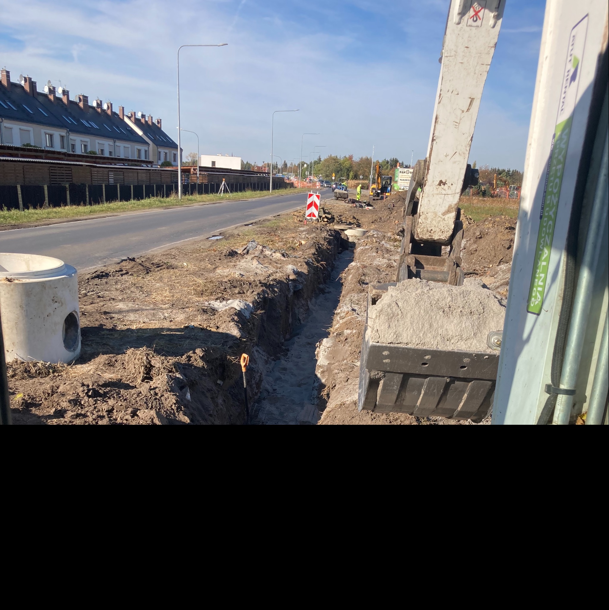 Wąski wykop liniowy wzdłuż drogi, z betonową studzienką obok i koparką w tle, robotnicy w odblaskowych ubraniach widoczni w oddali, znak drogowy ostrzegawczy.