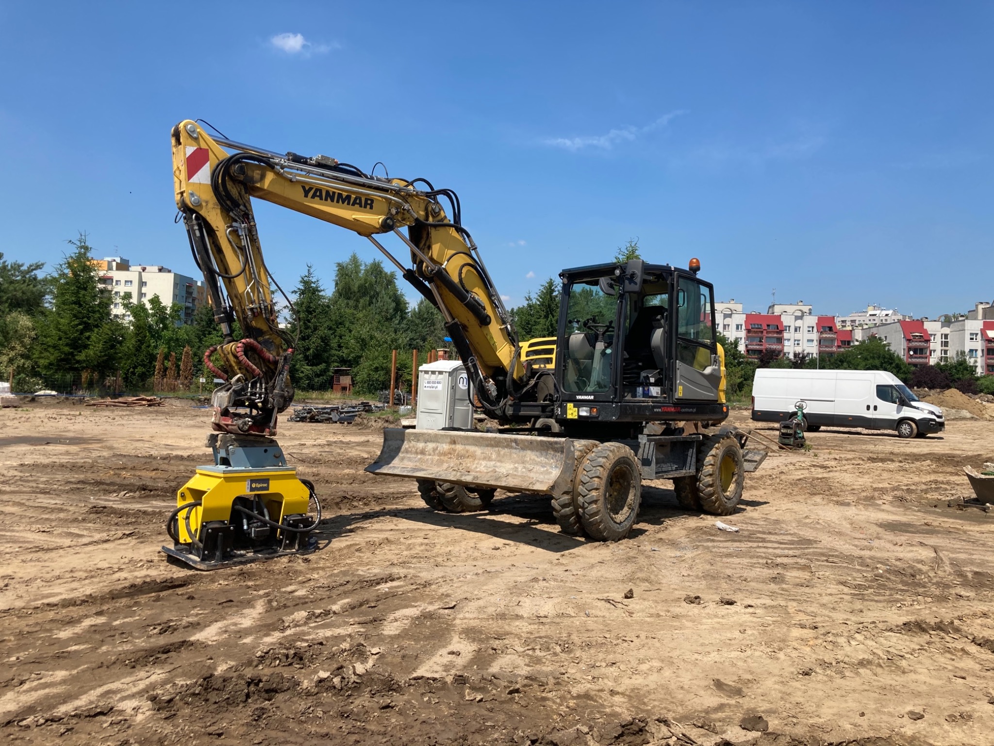 Żółta koparka Yanmar z wibro płytą na placu budowy, przygotowująca teren pod inwestycję, widoczna przenośna toaleta TOI-TOI i budynki mieszkalne w tle.