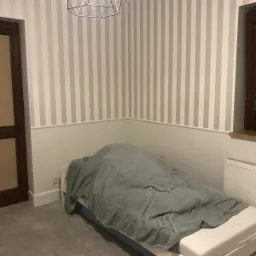 Pokój po odświeżeniu: Ściany w szaro-białe pasy, biała lamperia, łóżko przykryte szarym kocem, nowoczesna lampa geometryczna.