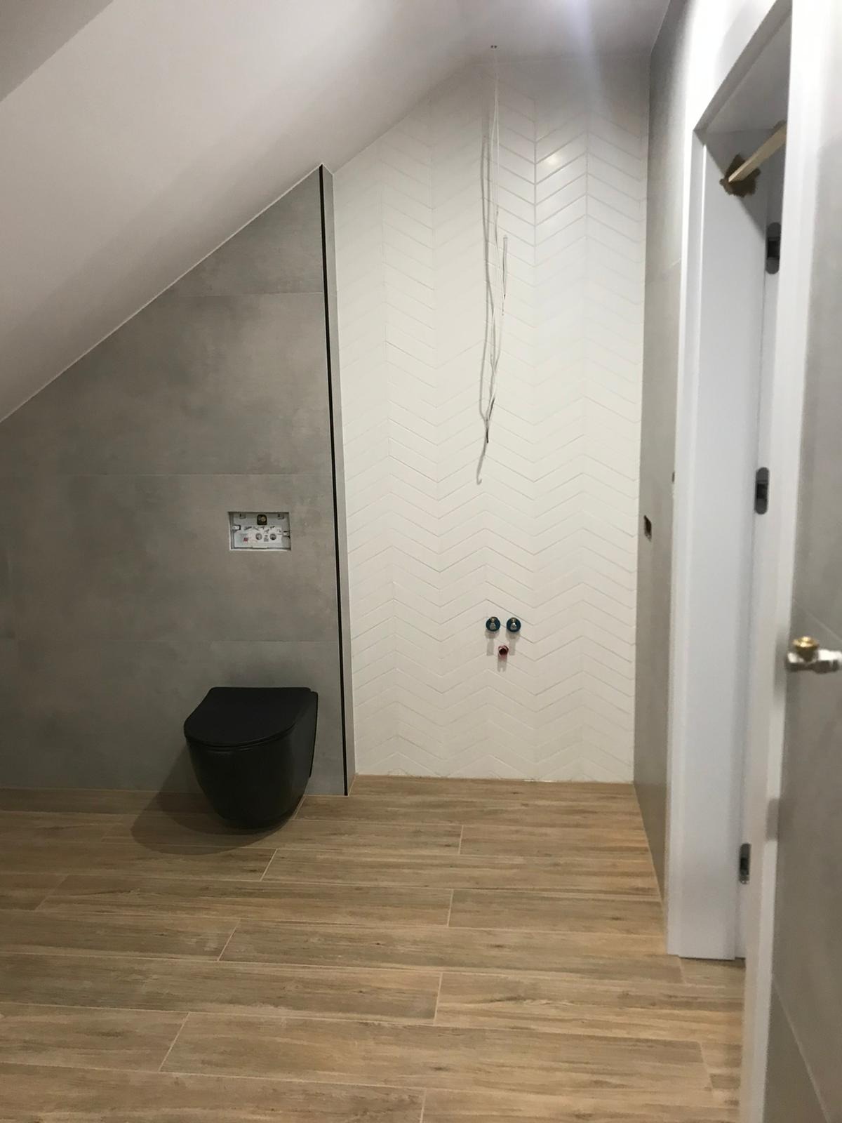 Ściana łazienki wyłożona białą chevronową mozaiką, z wiszącą czarną miską WC, drewnopodobne płytki podłogowe i wystające kable instalacji elektrycznej.