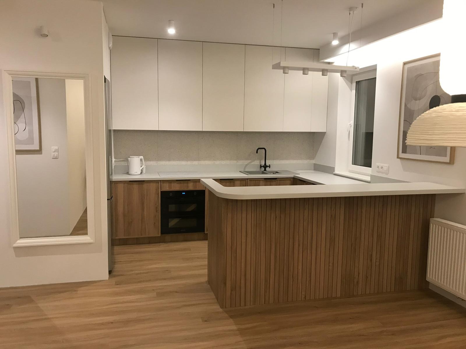 Nowoczesna kuchnia z białymi szafkami górnymi i drewnianymi dolnymi, wyspa z drewnianymi lamelami, jasna podłoga, czarna armatura i minimalistyczne oświetlenie.