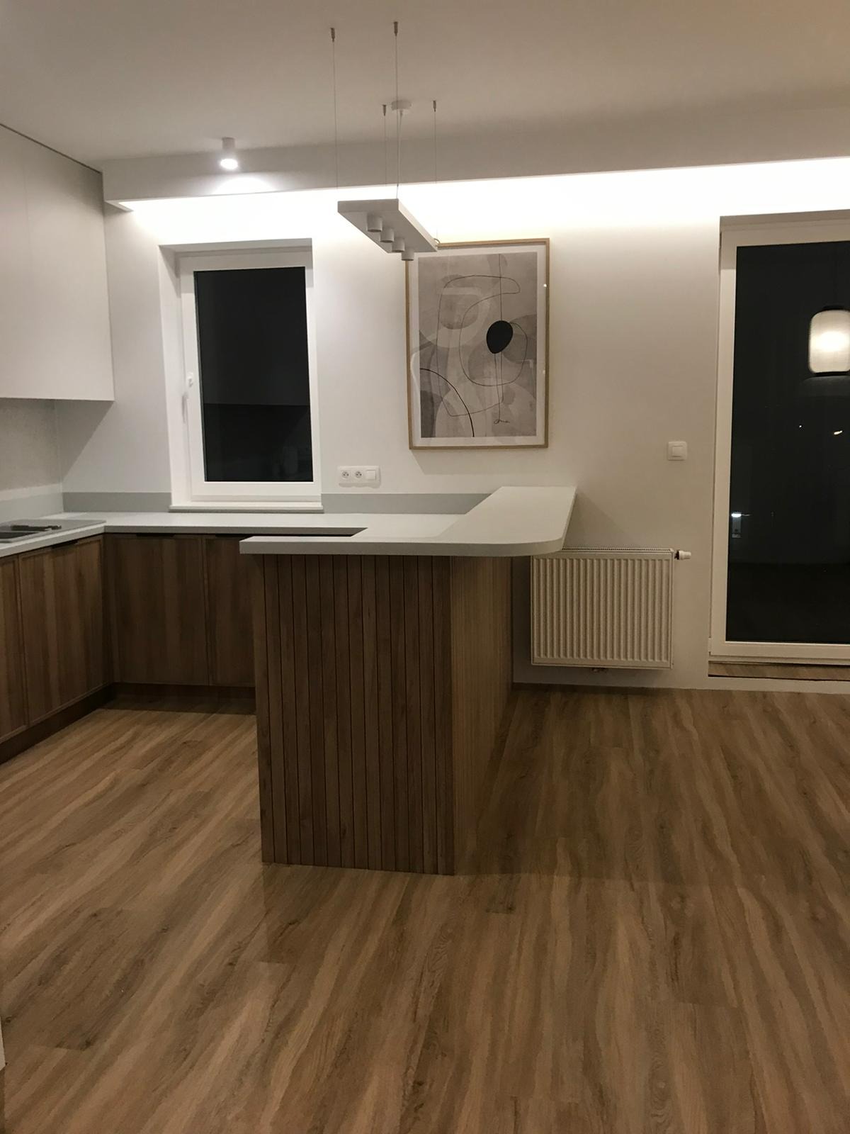 Nowoczesna kuchnia z drewnianymi szafkami i wyspą, jasny blat, drewniana podłoga, minimalistyczny obraz na ścianie, oświetlenie sufitowe.