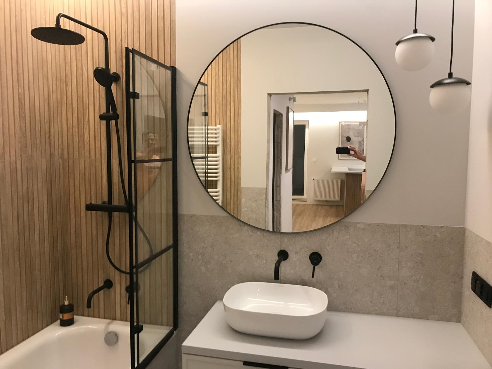 Nowoczesna łazienka z drewnianymi panelami ściennymi, czarną armaturą, okrągłym lustrem odbijającym wnętrze, białą umywalką nablatową i wiszącymi lampami kulami.