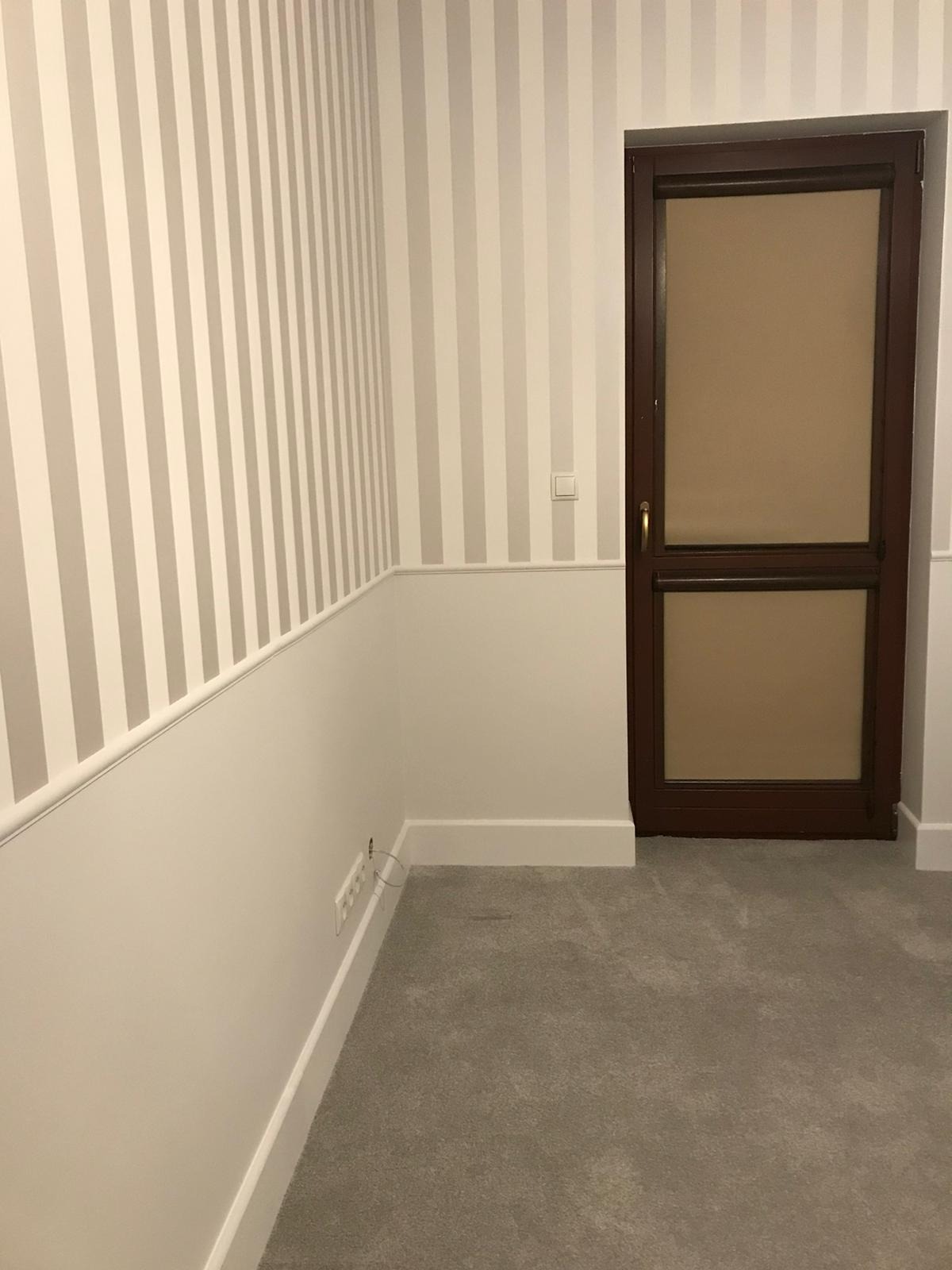 Pokój po remoncie: ściany w pionowe pasy beżu i bieli, biała lamperia, szara wykładzina, brązowe drzwi z szybą.
