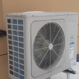 Zewnętrzna jednostka klimatyzacji typu split z widoczną kratką wentylacyjną i naklejką informacyjną, zamontowana na metalowym stelażu przy ścianie budynku, lekko przyprószona śniegiem.