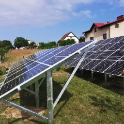 Instalacja paneli fotowoltaicznych na gruncie, widoczne panele pod kątem, domy w tle, zielona trawa i drzewa.