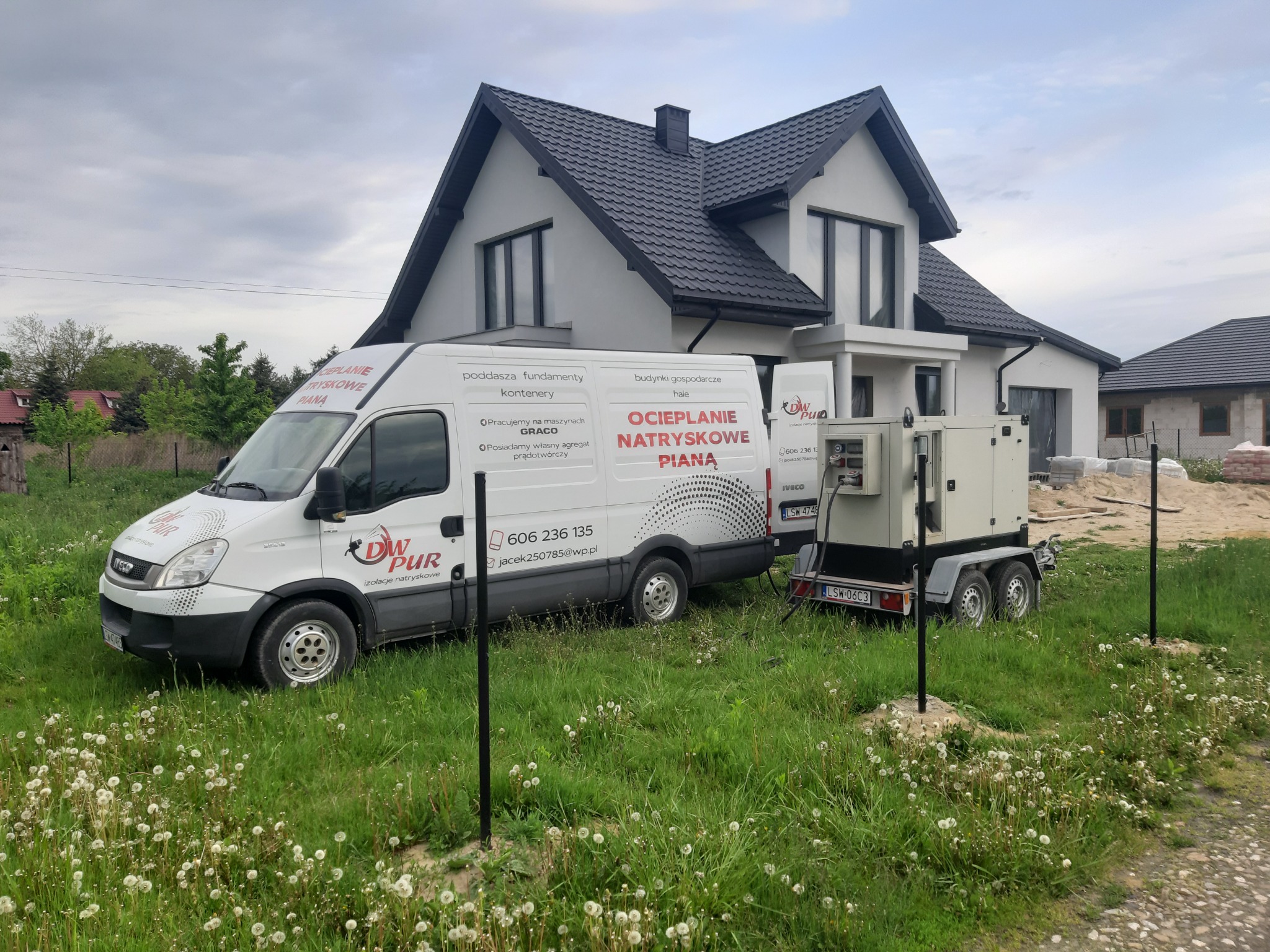 Biały samochód dostawczy z logo firmy i napisem 'Ocieplanie natryskowe pianą' zaparkowany na trawniku przed nowoczesnym domem jednorodzinnym z szarą dachówką, obok przyczepa z agregatem prądotwórczym.