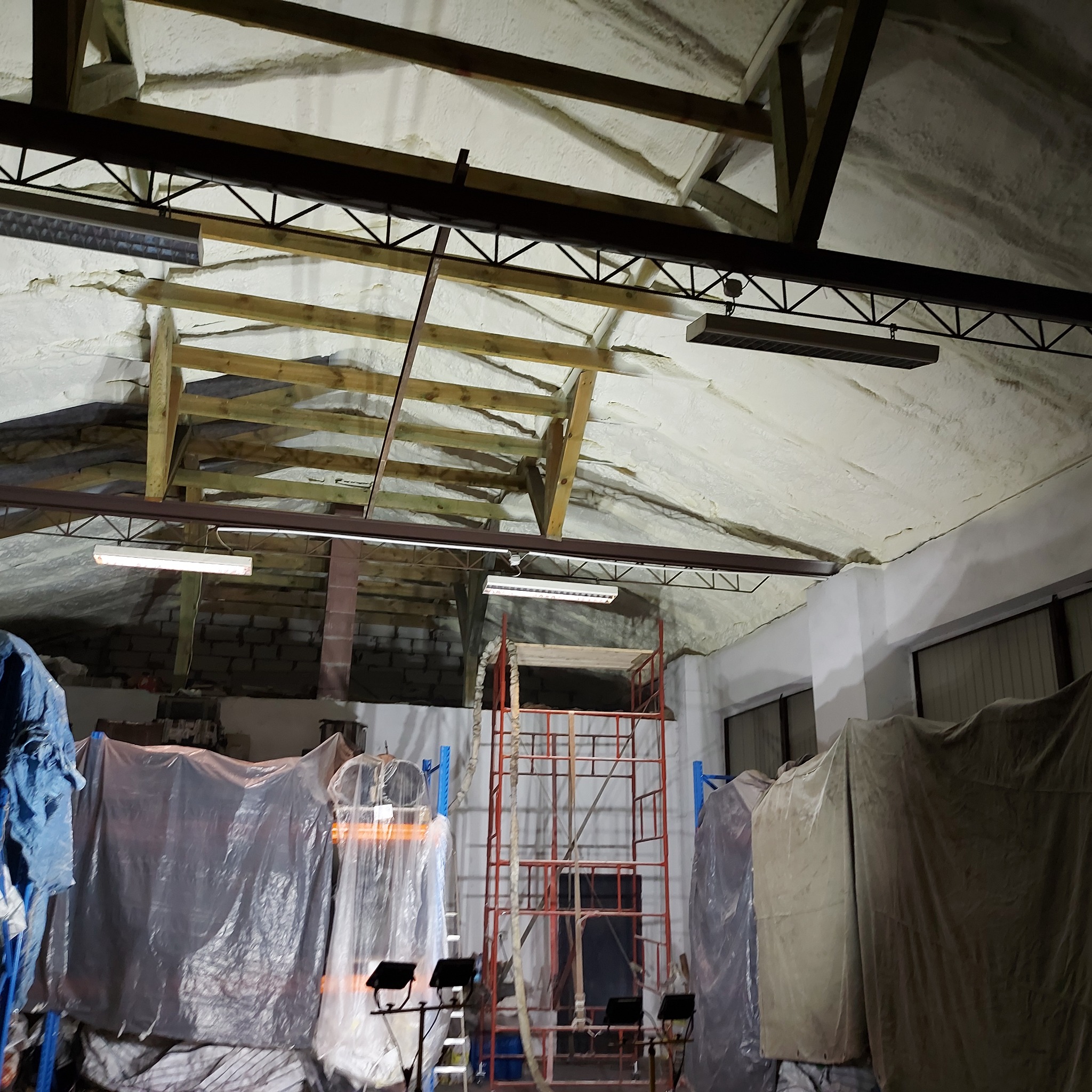 Poddasze po ociepleniu pianką PUR, widoczne drewniane elementy konstrukcyjne, lampy oświetleniowe i rusztowanie w pomieszczeniu.