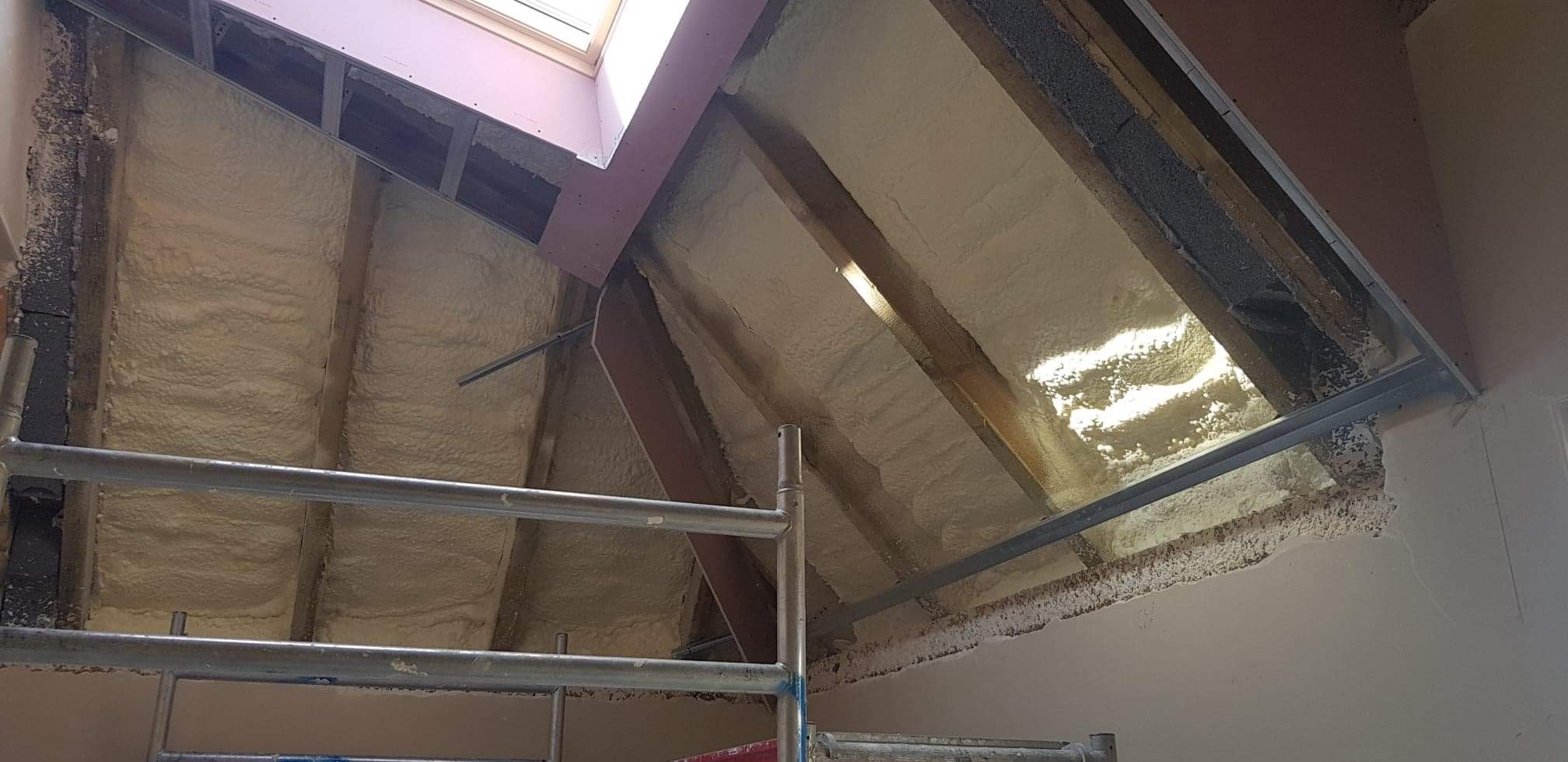 Poddasze w trakcie izolacji pianką poliuretanową, widoczne krokwie, okno dachowe, fragment rusztowania i nierównomiernie nałożona warstwa piany na skośnych powierzchniach.