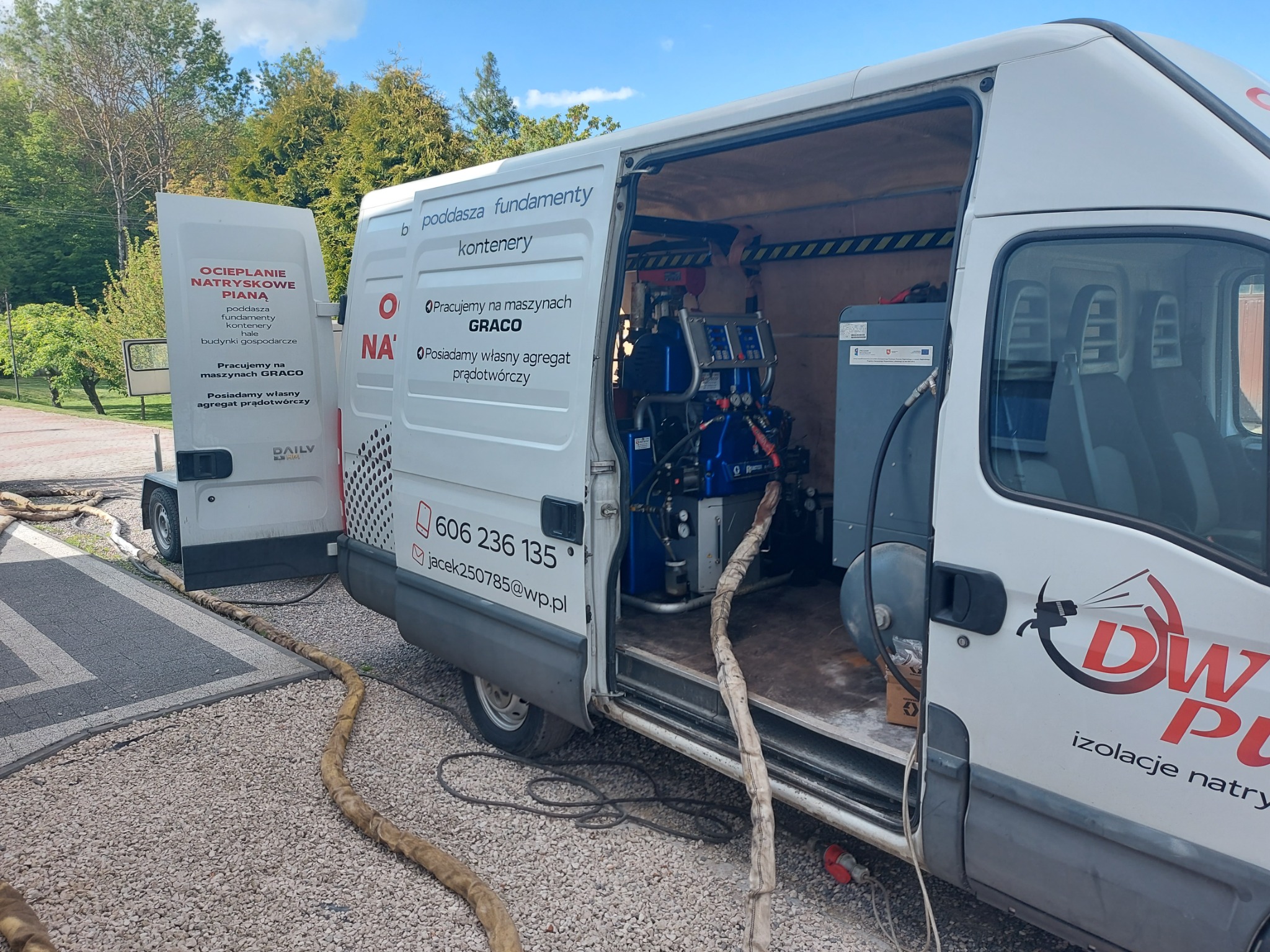 Otwarty samochód dostawczy z agregatem do natrysku pianki, widoczne węże, reklama firmy na karoserii, teren zielony w tle.