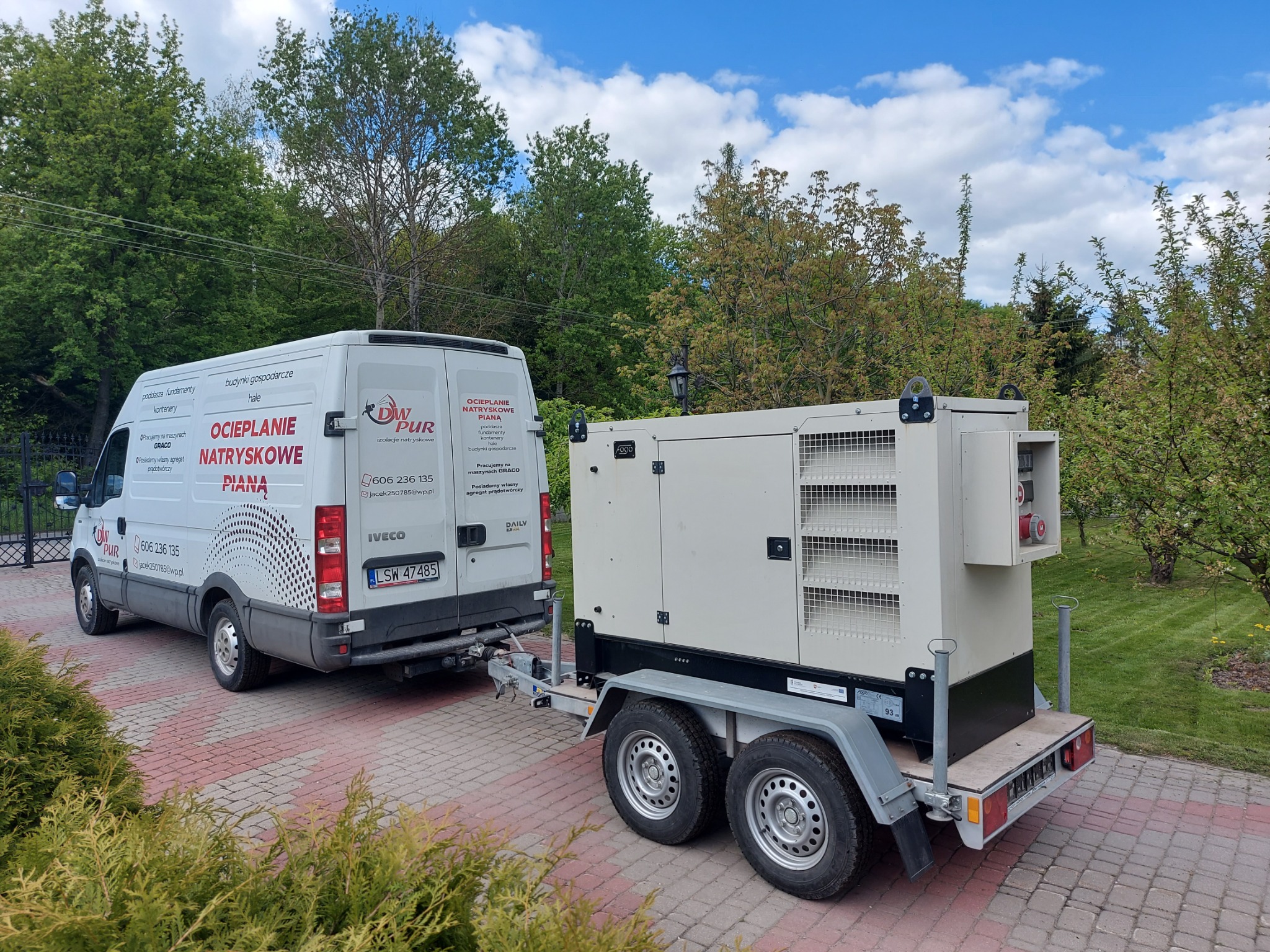 Bus firmy ocieplającej pianką PUR z przyczepą zawierającą agregat prądotwórczy, zaparkowany na brukowanym podjeździe przed posesją z zielonymi drzewami w tle.