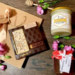 Kompozycja prezentowa: czekolada Cortez z migdałami, miód faceliowy w słoiku z etykietą Pasieka Tomala, słoik konfitury piernikowej z kokardą, drewniana nabierka do miodu i różowe goździki...