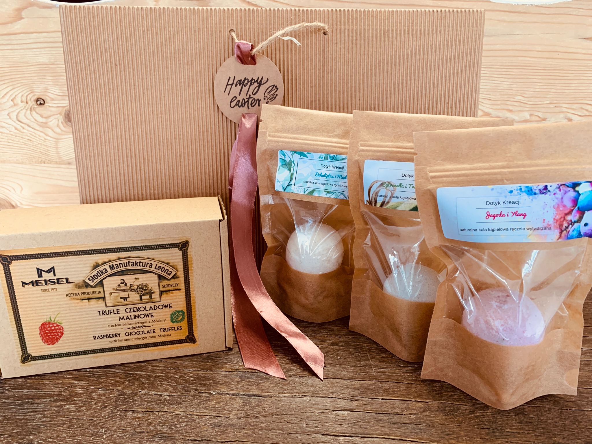 Zestaw prezentowy z tekturowym pudełkiem, czekoladowymi truflami malinowymi i kulami do kąpieli w ekologicznych opakowaniach, ozdobiony wstążką i zawieszką z napisem 'Happy Easter'.