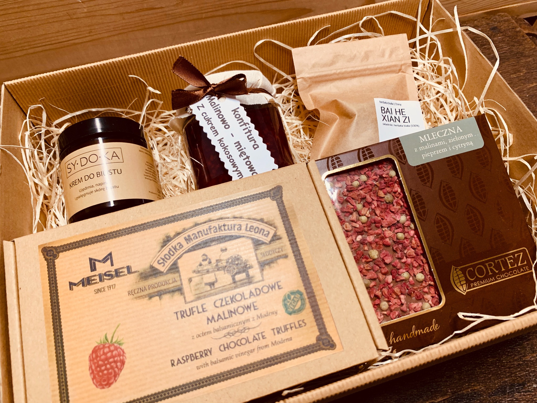Krem do biustu, konfitura malinowo-miętowa, herbata biała, czekolada z malinami i pieprzem w kartonowym pudełku wypełnionym siankiem dekoracyjnym, tworzące zestaw prezentowy.