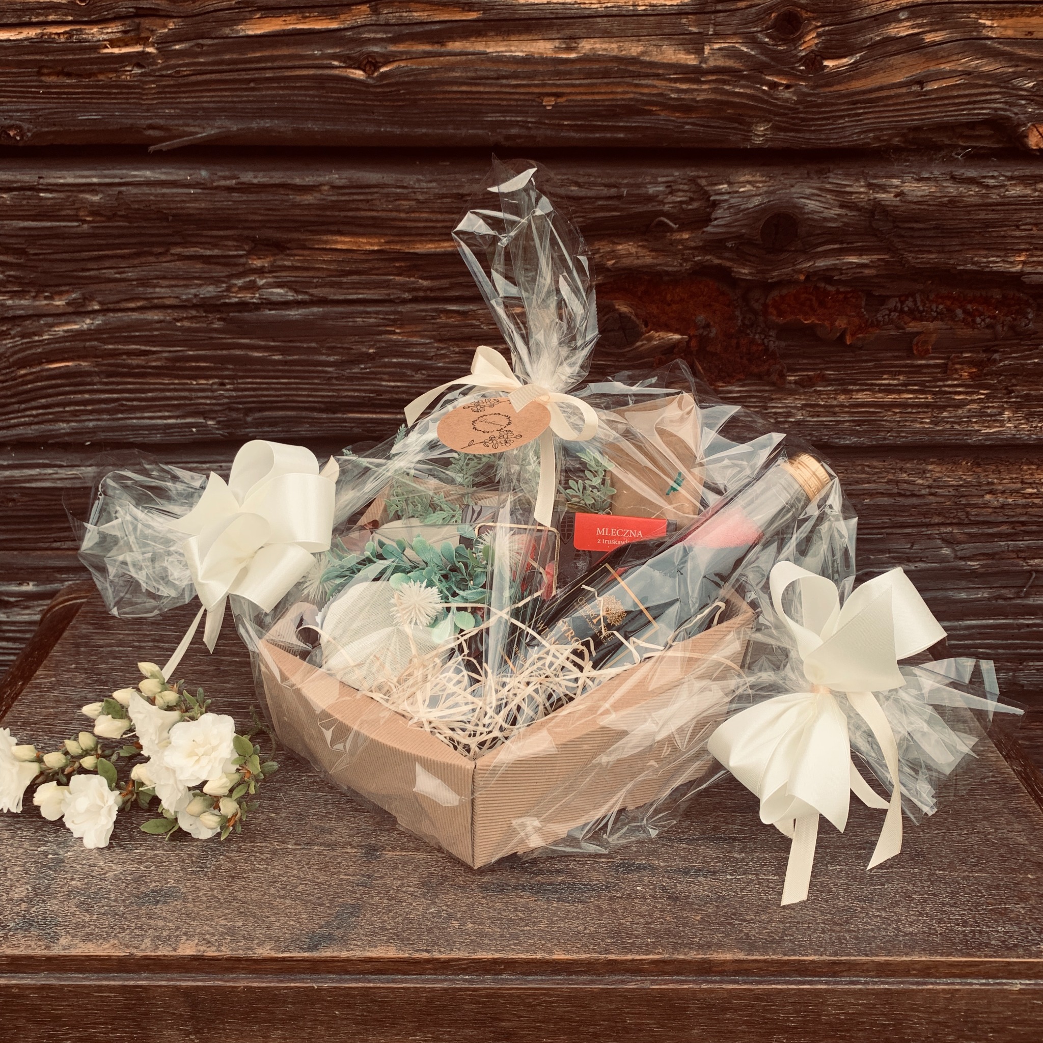 Kartonowy kosz z winem, słodyczami i dekoracjami, owinięty folią i ozdobiony kremowymi wstążkami, leżący na drewnianym blacie na tle rustykalnej drewnianej ściany.